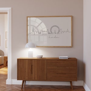 May include: A wooden dresser with three drawers and two cabinet doors. The dresser has a light brown finish and is sitting on a hardwood floor. A white lamp and a white bowl are on top of the dresser. A framed print of a line drawing of the London skyline is hanging on the wall behind the dresser.
