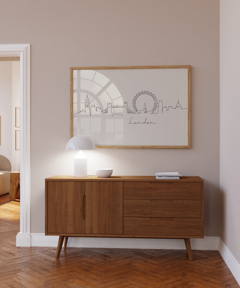 May include: A wooden dresser with three drawers and two cabinet doors. The dresser has a light brown finish and is sitting on a hardwood floor. A white lamp and a white bowl are on top of the dresser. A framed print of a line drawing of the London skyline is hanging on the wall behind the dresser.