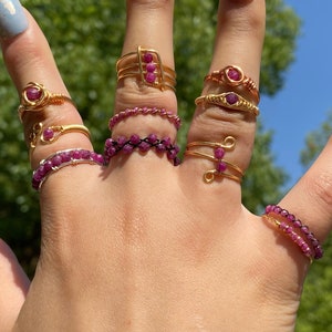 Puede incluir: Una mano muestra una colección de anillos hechos a mano. Los anillos presentan piedras preciosas de rubí y están elaborados con alambre de oro y plata. Los diseños incluyen alambre envuelto, bandas de cuentas y un anillo en forma de rosa. El fondo es una vista borrosa de follaje verde y cielo azul.