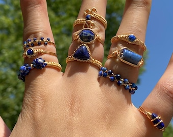 May include: A hand wearing a set of seven gold rings with blue lapis lazuli stones. The rings are all different styles, some with a single stone and others with multiple stones.