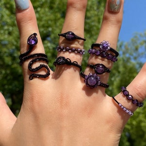 Könnte beinhalten: Eine Hand trägt mehrere Ringe mit Amethyst-Edelsteinen. Die Ringe sind aus schwarzem Draht gefertigt und haben verschiedene Designs, darunter eine Schlange, eine Blume und ein schlichtes Band.