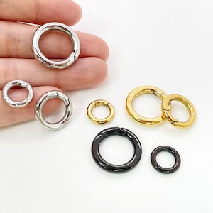 Puede incluir: Una colección de anillos redondos de cierre de resorte metálicos en varios tamaños y acabados. Los cierres son plateados, dorados y negros. Los cierres están abiertos, mostrando el mecanismo de resorte. Para la fabricación de joyas y manualidades.