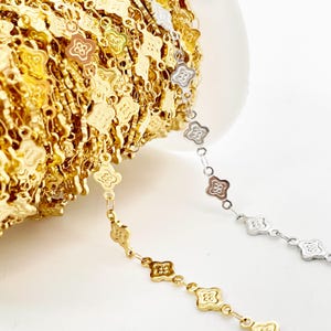 Puede incluir: Primer plano de un carrete de cadena dorada con un diseño en forma de trébol. Se ve un hilo separado de cadena plateada con el mismo diseño de trébol. Las cadenas están sobre un fondo blanco.