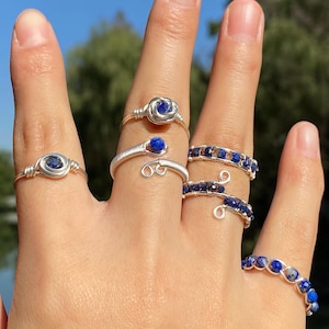 以下が含まれることがあります： 青い宝石のアクセントが付いた銀の指輪を5つつけた手。指輪はワイヤー製で、バラ、渦巻き、小さな石のバンドなど、さまざまなデザインが施されています。