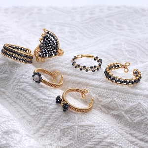 May include: A selection of handmade wire rings. The rings are made with gold-coloured metal and black beads. Designs include a butterfly shape, floral accents, and beaded bands. The rings are displayed on a white, textured surface.