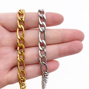 Può includere: Due catene curb, una dorata e una argentata, entrambe con un designo a maglia piatta di forma ovale. Le catene sono tenute in una mano, con la catena dorata a sinistra e la catena argentata a destra.