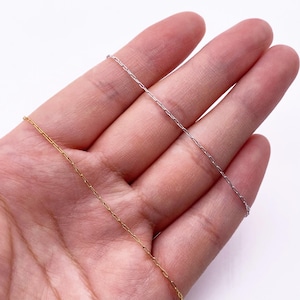 Peut inclure: Gros plan de deux fines chaînes de collier. L'une est dorée, l'autre argentée. Les chaînes sont fines et présentent un design à maillons rectangulaires. Les chaînes sont présentées sur un fond clair.