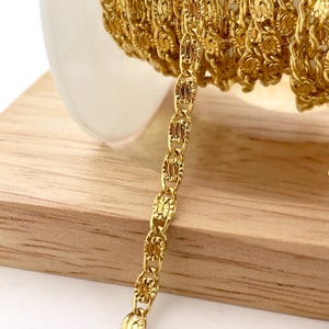 Puede incluir: Primer plano de una cadena dorada con un diseño de eslabones ovalados. La cadena está enrollada sobre una superficie blanca y un bloque de madera. La cadena es de un color dorado brillante.