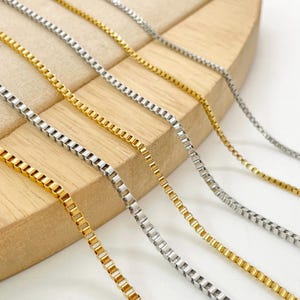 Puede incluir: Una colección de cuatro cadenas de eslabones cuadrados dispuestas en paralelo, que incluye dos cadenas de tono dorado y dos de tono plateado. Cada cadena está formada por pequeños eslabones metálicos cuadrados que crean un diseño lineal flexible.