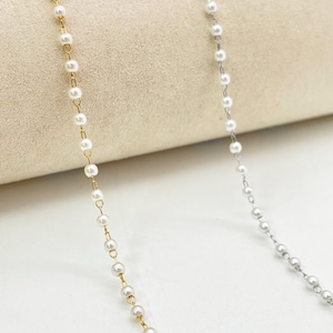 Puede incluir: Primer plano de dos delicados collares. Uno es dorado, el otro plateado, ambos adornados con pequeñas perlas blancas redondas. Los collares se exhiben sobre un fondo neutro, blanquecino, resaltando su elegante sencillez.
