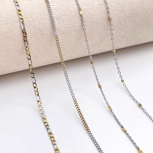 May include: Close-up of four necklaces in silver and gold. The first features a figaro chain, the second a curb chain. The third and fourth have delicate chains with gold details. The chains are displayed against a neutral background.