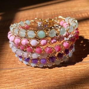 Könnte beinhalten: Ein mehrlagiges Armband mit verschiedenen Edelsteinperlen, darunter grün, rosa, rot, lila und weiß. Die Perlen sind auf goldenen Draht gefädelt und das Armband ist für das Tragen am Handgelenk konzipiert.