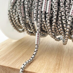 May include: Close-up of a spool of shiny, silver-colored chain. The chain is made of small, round beads and rectangular links, creating a textured appearance. The chain is coiled on a white spool, resting on a light-colored wooden surface.