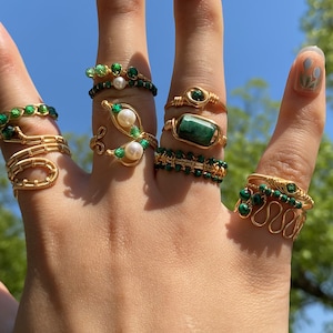 Puede incluir: Un maniquí de mano con varios anillos de oro con piedras verdes y blancas. Los anillos están hechos de alambre y presentan diferentes diseños, incluyendo una forma de vaina de guisante, una piedra rectangular y una serie de pequeñas piedras verdes.