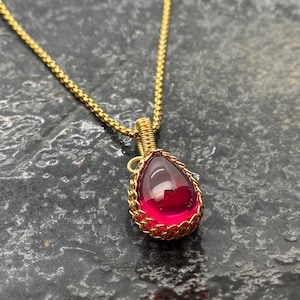 Op de afbeelding: Een gouden ketting met een traanvormige hanger. De hanger is gemaakt van gouden draad en heeft een grote, felroze edelsteen.