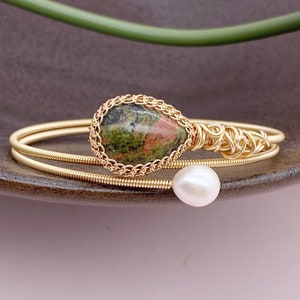 Könnte beinhalten: Ein goldfarbenes, drahtgewickeltes Armband mit einem grün-roten Edelstein und einer weißen Perle. Das Armband besteht aus zwei Drähten.