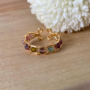 Puede incluir: Un anillo de alambre dorado con piedras preciosas de colores. El anillo está hecho con un diseño delicado de alambre y presenta una variedad de pequeñas piedras preciosas redondas en tonos de rosa, morado, verde y amarillo.