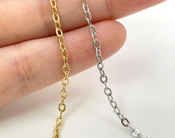 Peut inclure: Deux chaînes de bijoux délicates, l'une dans un ton or brillant et l'autre dans un ton argenté, présentant de petits maillons ovales avec une surface texturée et facettée qui capte la lumière. Les chaînes sont présentées côte à côte.