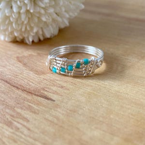 Puede incluir: Un anillo de alambre plateado con cuentas de color azul turquesa. El anillo está tejido con varios hilos de alambre.