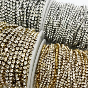 Puede incluir: Primer plano de tres carretes de cadena de strass. La cadena está hecha de pequeños strass transparentes que están conectados por una base metálica. Los carretes son blancos y tienen una cadena dorada, plateada y transparente.