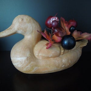 Puede incluir: Un macetero de cerámica con forma de pato con un esmalte marrón claro. El macetero está decorado con un racimo de bayas rojas y negras y hojas de otoño.