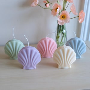 Può includere: Cinque candele a forma di conchiglia in diversi colori pastello: verde, viola, rosa, crema e blu. Le candele sono disposte su una superficie in legno chiaro.