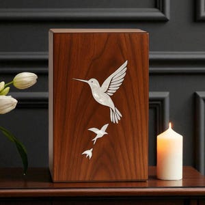May include: A wooden urn with a hummingbird design. The rectangular urn has a dark wood finish. A white hummingbird is depicted in flight, with two smaller birds below. A lit candle and flowers are on the table.