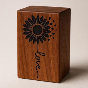 Op de afbeelding: Een rechthoekig houten blok met een zwart zonnebloemontwerp en het woord "love" gegraveerd op de voorkant. Het blok heeft een warme bruine kleur. Kleine hartvormen zijn verspreid rond de zonnebloem.