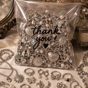 Peut inclure: Un sachet en plastique transparent rempli de bijoux argentés assortis, incluant des chaînes, des breloques en forme de cœur, des pendentifs papillon et des perles, avec le texte "thank you !" imprimé en noir. Des bagues et breloques sont disposées devant.