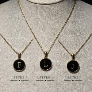 Puede incluir: Tres collares dorados con colgantes circulares con centro de esmalte negro y letras F, L y J doradas. Cada colgante cuelga de una delicada cadena dorada. Debajo de cada collar, el texto dice LETTRE F, LETTRE L y LETTRE J.