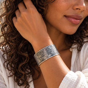 Peut inclure: Un large bracelet manchette en argent orné d'un motif floral en relief complexe avec des détails oxydés dans les zones en creux. La surface métallique présente une finition polie sur les bords, soulignant les gravures botaniques détaillées.