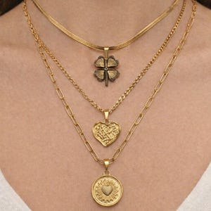 以下が含まれることがあります： 3つの異なるペンダントが付いた、重ね着けされたゴールドネックレスセット。一番上はクローバーチャーム、真ん中はハート、一番下はハートのデザインの丸いペンダント。チェーンはスタイルが異なり、ジュエリーに視覚的な面白さを加えています。