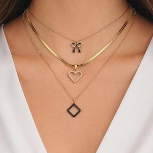 以下が含まれることがあります： 4つのゴールドチェーンが特徴のレイヤードネックレスセット。一番上のチェーンには小さなリボンペンダント、2番目はヘリンボーンチェーン、3番目はハートペンダント、一番下は正方形のペンダントが付いています。