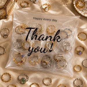 Könnte beinhalten: Ein durchsichtiger Plastikbeutel mit mehreren Metallringen, auf dessen Vorderseite der Text "Happy every day" und "Thank you!" aufgedruckt ist. Der Beutel ist von verschiedenen gold- und silberfarbenen Ringen mit unterschiedlichen Designs umgeben.