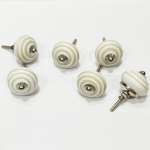 May include: Set of six white ceramic cabinet knobs with beige stripes and silver accents. The knobs have a round shape and are designed for use on cabinets, drawers, or furniture.
