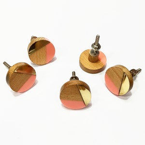 Könnte beinhalten: Fünf runde Holzknauf für Schränke mit geometrischem Design in Pink und Gold. Die Knöpfe haben eine silberne Schraube zum Befestigen an einer Schranktür.