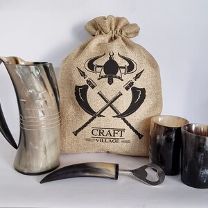 Può includere: Un set di tre corni da bere, un apribottiglie con manico in corno e una borsa di juta con un design vichingo. La borsa ha la scritta "CRAFT VILLAGE" stampata.