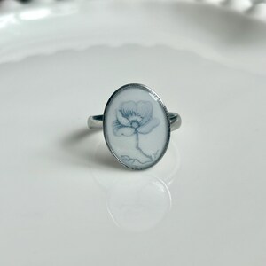 May include: A silver-toned ring featuring an oval-shaped setting with a white background. The center of the oval displays a delicate, light blue line drawing of a single flower with petals and a stem, encased under a smooth, clear surface.