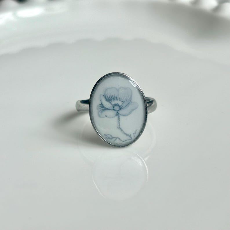 May include: A silver-toned ring featuring an oval-shaped setting with a white background. The center of the oval displays a delicate, light blue line drawing of a single flower with petals and a stem, encased under a smooth, clear surface.