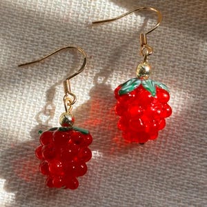 May include: A pair of gold dangle earrings with red glass grapes. The grapes are clustered together and have a green leaf on top.