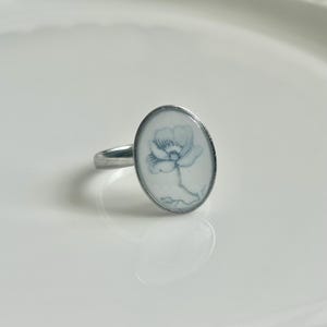May include: A silver-toned ring featuring an oval-shaped setting with a white background and a delicate, light blue line drawing of a single flower. The metal band is smooth and polished, showcasing a simple design with a floral motif.