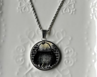 May include: A silver-toned rope chain necklace featuring a circular pendant. The pendant displays a black and white illustration of a cauldron sitting over a fire, set against a brick wall background, enclosed within a polished metal frame.