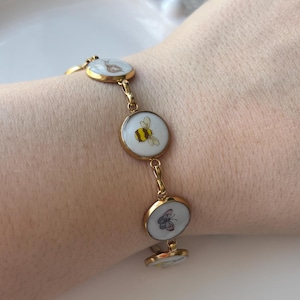 May include: Gold-toned bracelet featuring circular charms with insect and floral designs. The charms have white backgrounds with a bee, butterfly, and flower illustrations. The bracelet is connected by delicate gold links.