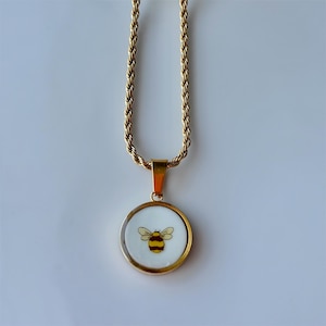 May include: Gold-toned necklace with a round pendant featuring a bee design. The bee is yellow and black, set against a white background. The chain has a rope-like texture.