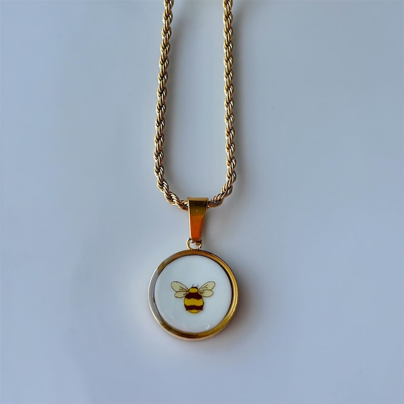 May include: Gold-toned necklace with a round pendant featuring a bee design. The bee is yellow and black, set against a white background. The chain has a rope-like texture.