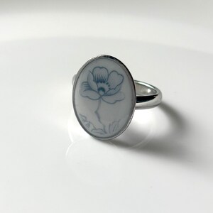 May include: A silver-toned ring featuring an oval-shaped white ceramic inlay. The center of the inlay displays a delicate, light blue line drawing of a single flower with petals and a stem, set within a polished metal band.