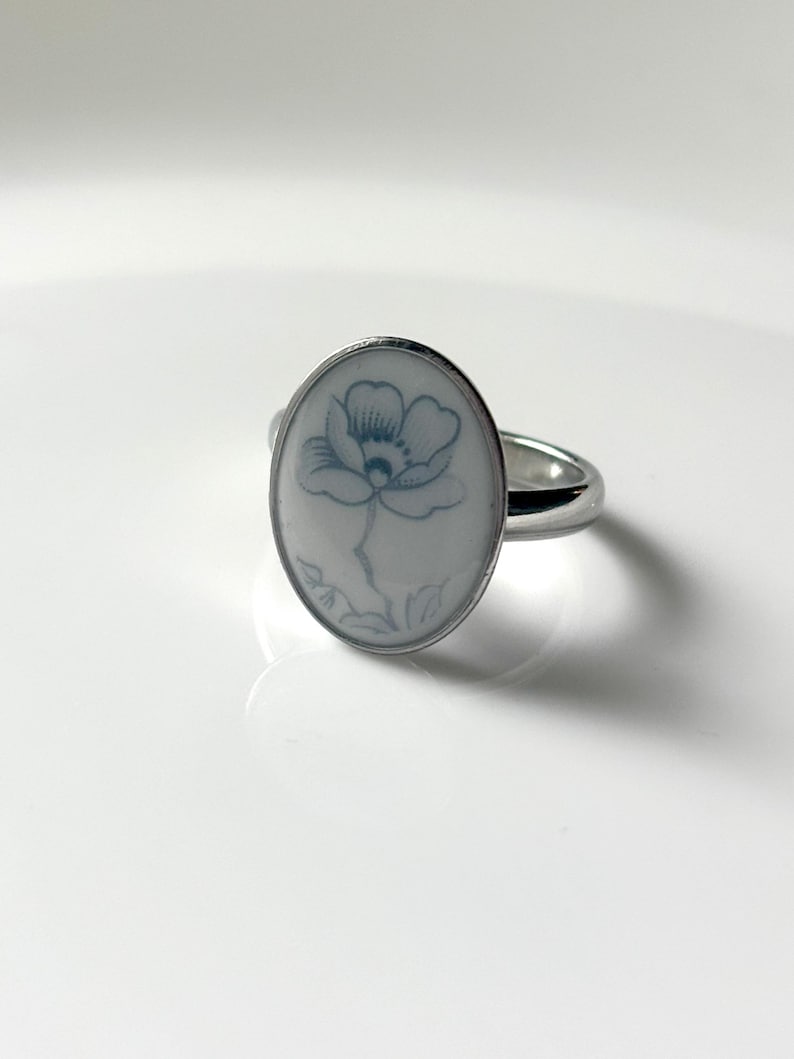 May include: A silver-toned ring featuring an oval-shaped white ceramic inlay. The center of the inlay displays a delicate, light blue line drawing of a single flower with petals and a stem, set within a polished metal band.