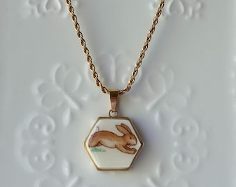 May include: Gold-toned necklace with a hexagonal pendant featuring a brown rabbit illustration on a white background. The rabbit is depicted in mid-run over a patch of green grass. The chain has a twisted rope design.
