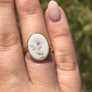 May include: A gold ring featuring an oval-shaped white inlay with a hand-painted illustration of a single pink flower with green leaves and a brown stem. The ring has a smooth, polished gold band that encircles the finger.