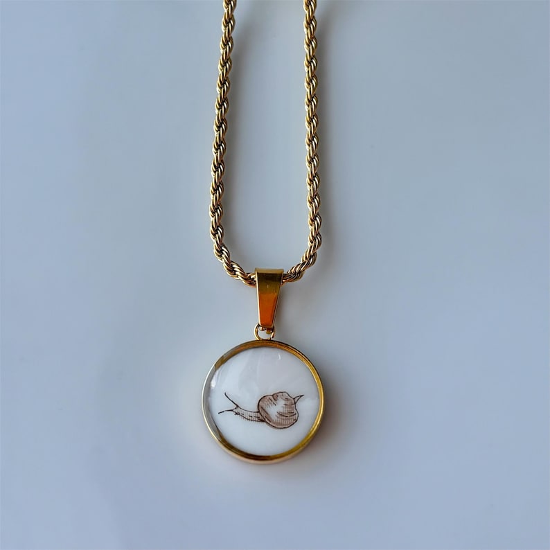 May include: Gold-toned necklace with a round pendant featuring a brown snail illustration on a white background. The pendant is encased in a gold frame and hangs from a gold rope chain.
